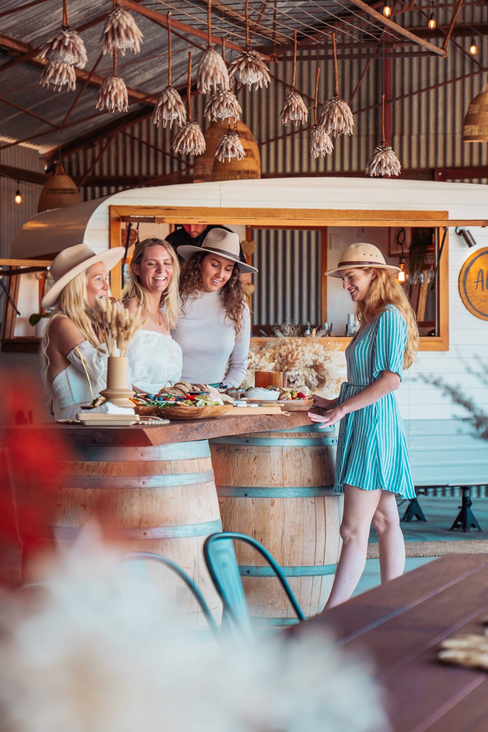 Four women share a joyful moment at a rustic, chic bar in Perth, Western Australia. They are dressed casually, with three of them wearing hats, and they're all smiling and engaging with each other over a wooden bar table adorned with a spread of delicious-looking food.