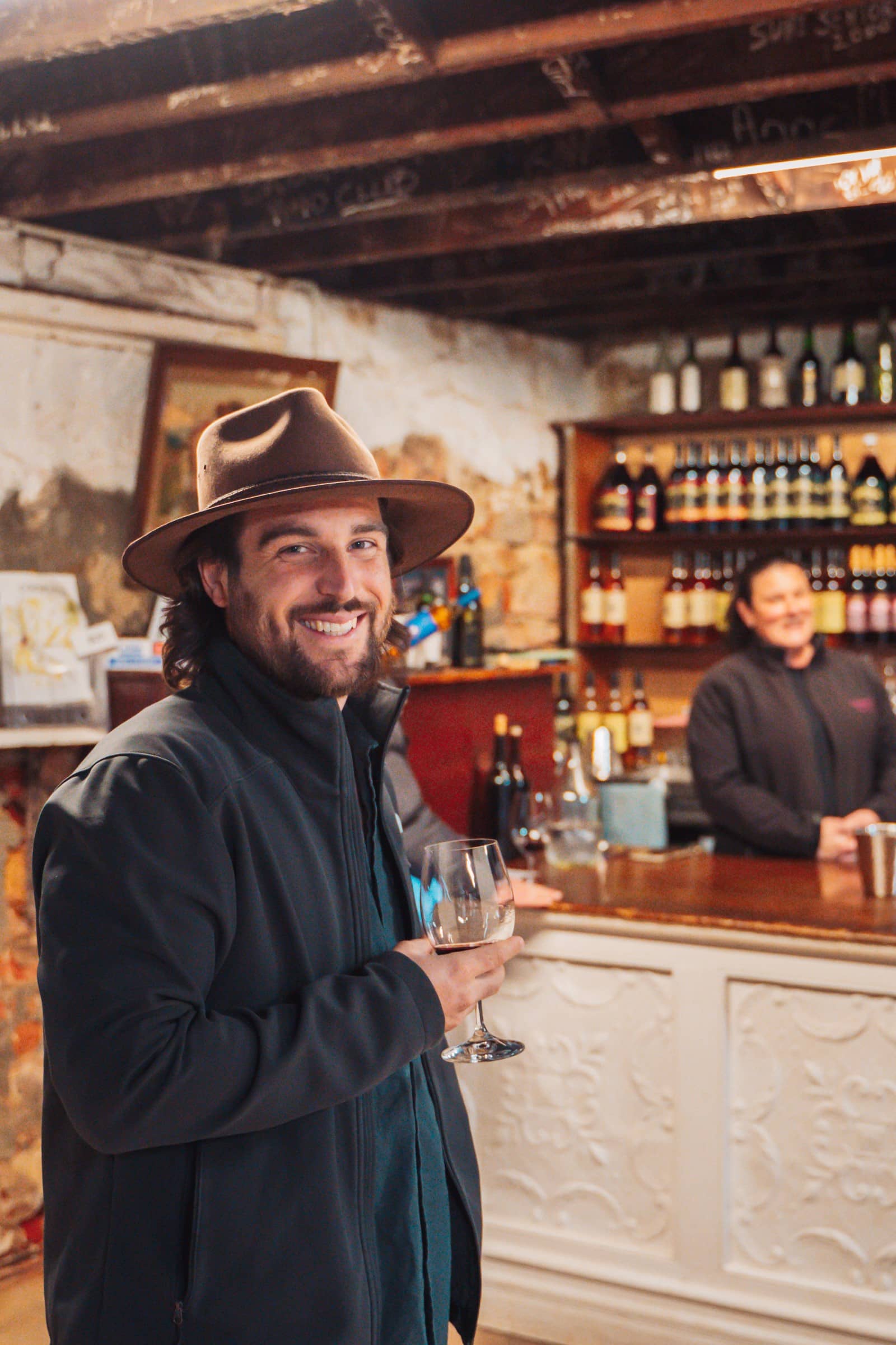 Chris of Salt and Charcoal hat and navy coat smiles while holding a wine glass, standing in a rustic bar with vintage charm in Perth, Western Australia. A bartender is visible in the background.