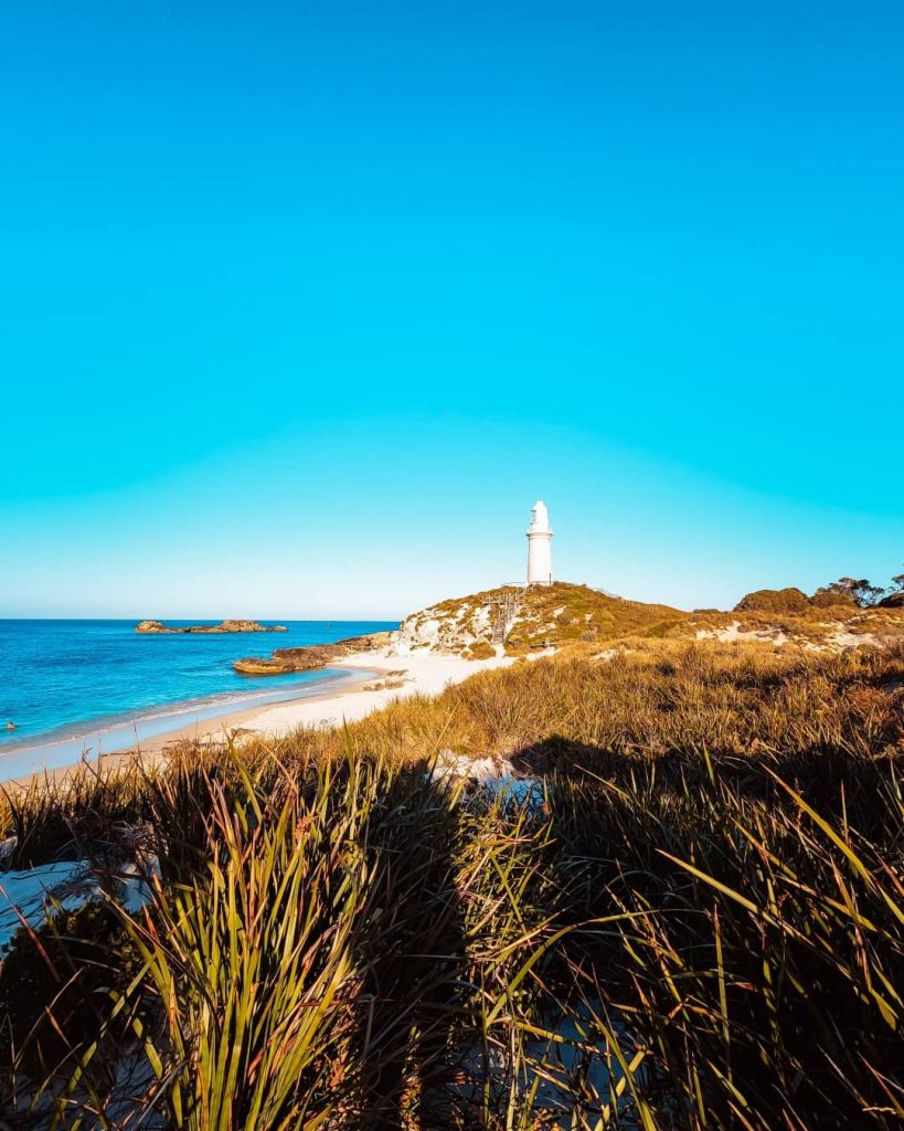 View of Bathurst Lighthouse on Rottnest Island, situated on a hill with sandy beaches and coastal vegetation in the foreground, under a clear blue sky.