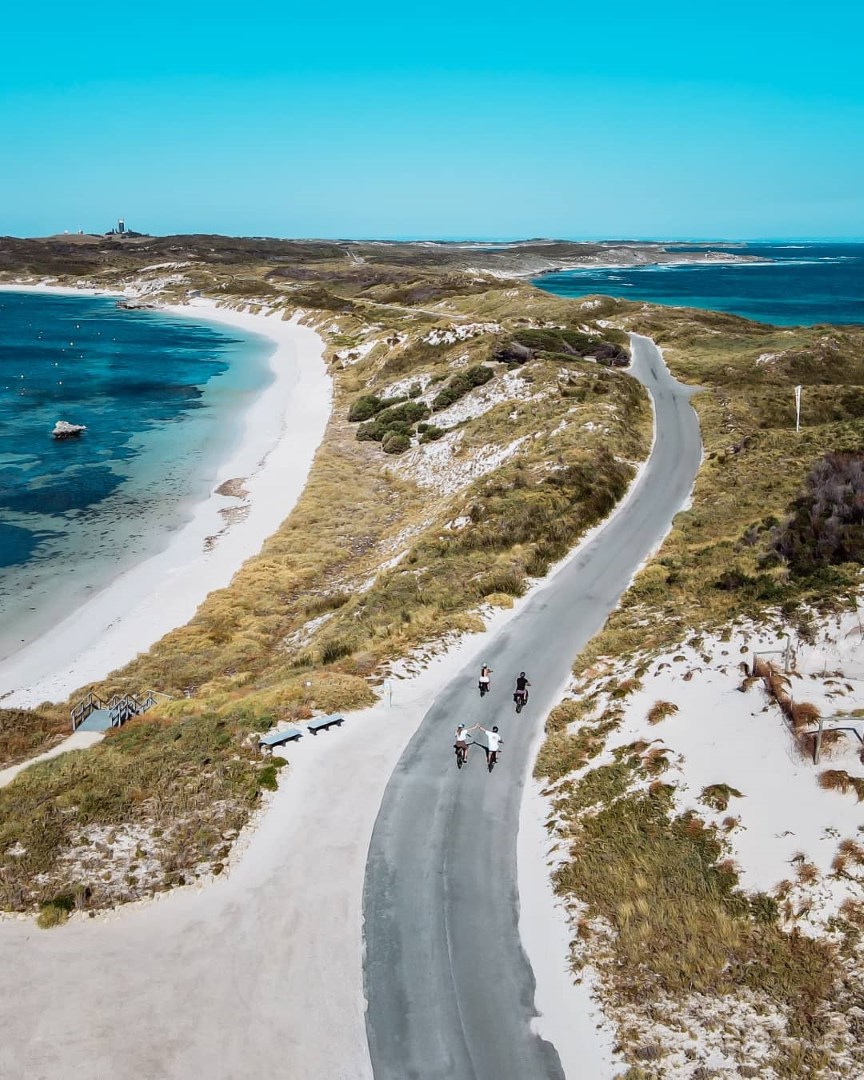 Aerial view of a coastal road on Rottnest Island with four cyclists riding along it. The road winds between sandy dunes and the blue ocean, with lush vegetation on either side.
