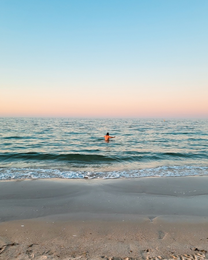 Chris swimming alone in the calm waters of the Busselton Foreshore, a cloudless sky that stretches out in soft shades of blue and pink. The shore is calm, just a gentle wash of waves touching the sandy beach.