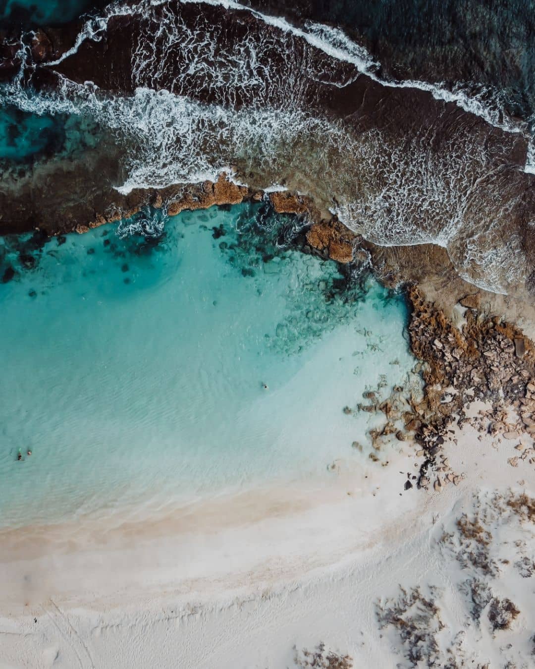 Aerial view of Cape Range National Park's coastline with clear turquoise waters meeting a rocky shoreline. Waves crash against the rocks, blending with a white sandy beach.
