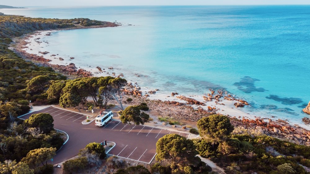 Aerial image showing the Eagle Bay area, one of the things to do in Dunsborough a town near Busselton. The image shows a tranquil beach with clear turquoise waters gently lapping against a rocky shoreline. Dense green foliage surrounds a parking area where only a vintage-style camper van is parked.