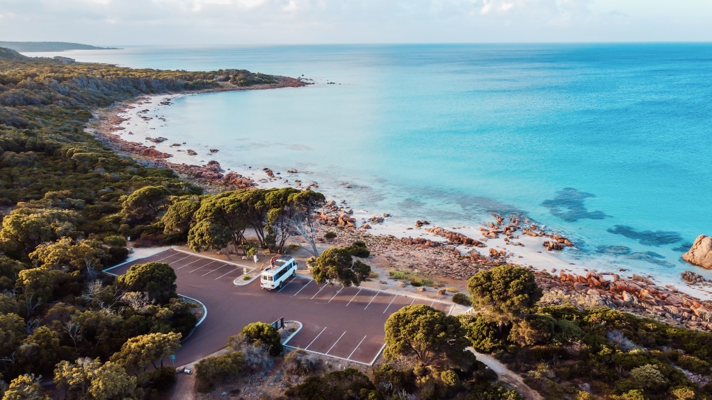 Aerial view of the Eagle Bay region in Dunsborough Western Australia showing a pristine coastline, turquoise waters, rocky shores, and dense greenery with a lone camper van parked in an empty parking lot near the shoreline.