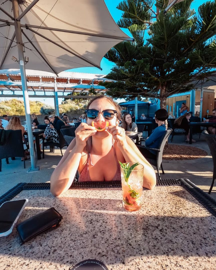 A woman sitting at an outdoor cafe table in Rottnest Island Western Australia, playfully holding a slice of fruit in front of her mouth like a smile.