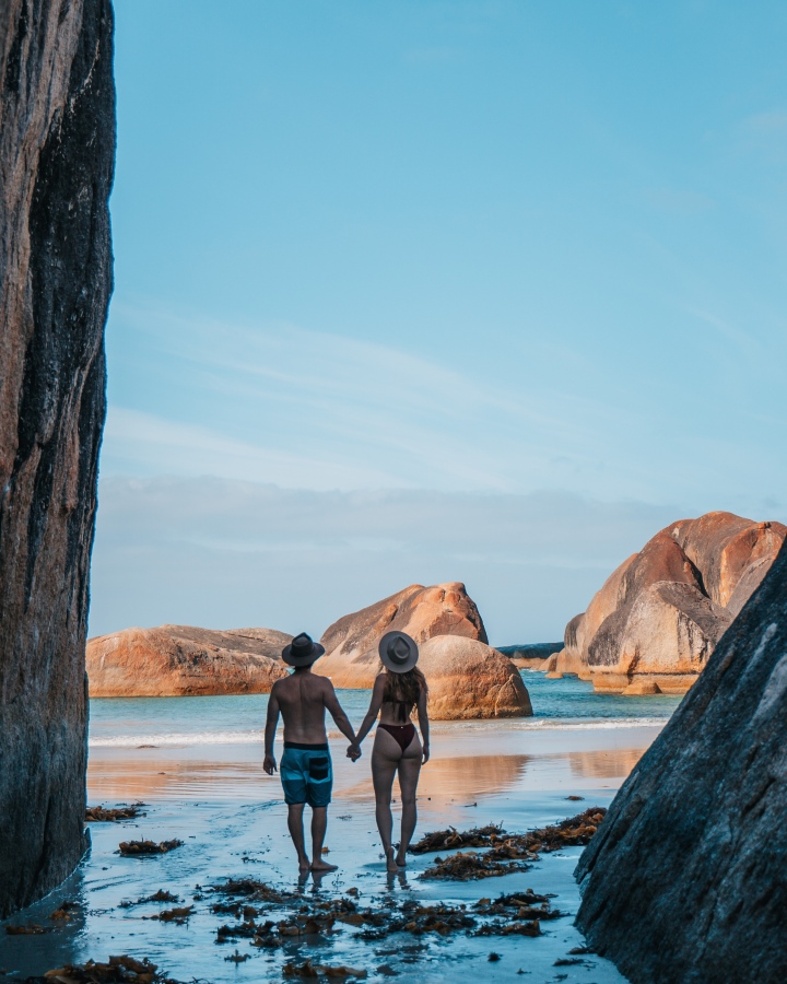 A couple holding hands while walking on the sandy beach of Elephant Rocks in Denmark, a town near Bremer Bay, in Western Australia. They are framed by large boulders as they face the calm, blue ocean in the distance, surrounded by more impressive rock formations.