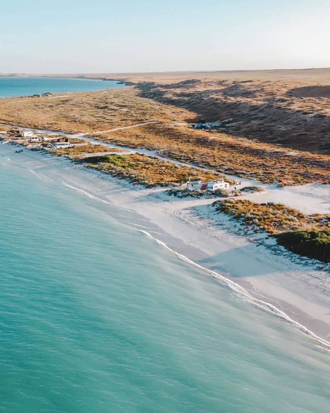 Aerial view of Cape Range National Park's coast, showing small buildings along a pristine white sandy beach with turquoise waters, sparse vegetation, and rugged terrain.





