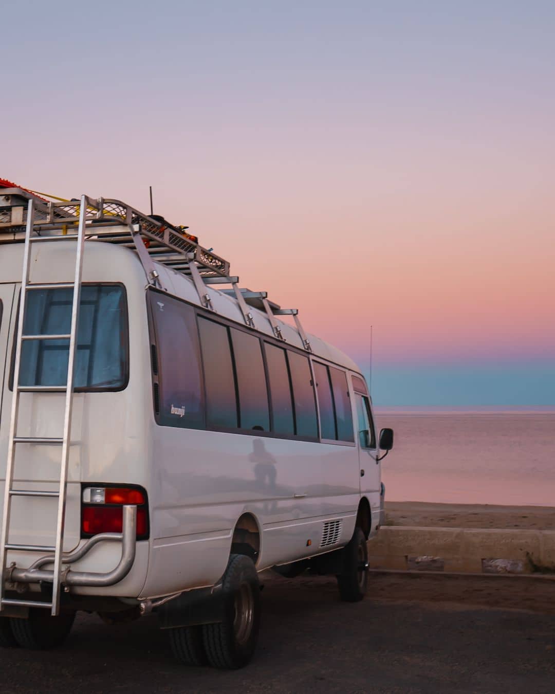 A white camper van parked near the beach at sunset, with the sky displaying soft pink and purple hues. The peaceful ocean and pastel-colored sky create a serene evening scene.