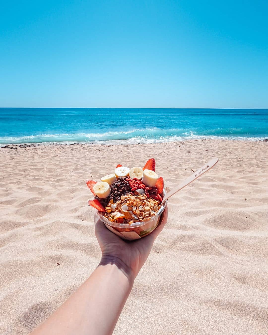 A hand holds a bowl of granola, fruit, and yogurt on a sandy beach with the ocean in the background, under a clear blue sky.




