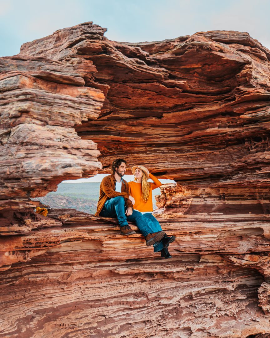 A couple sits within Nature's Window, a natural rock formation in Kalbarri National Park, Western Australia. They are framed by the layered red sandstone enjoying the scenic views.