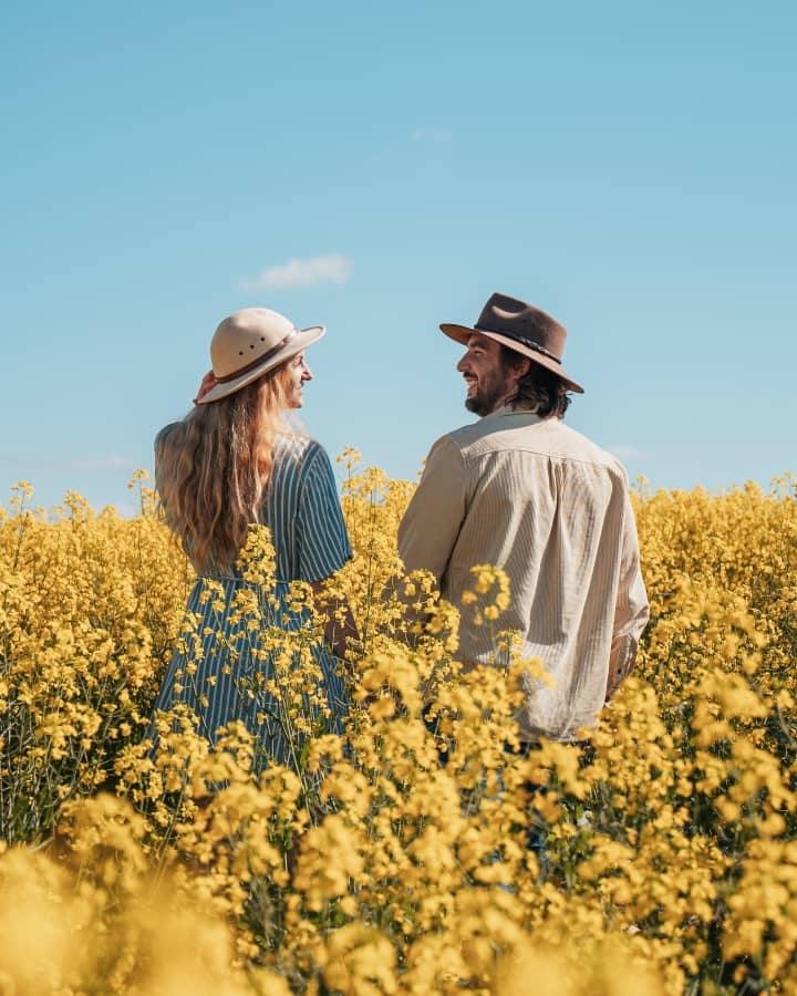A happy couple in hats enjoying a field of vibrant yellow wildflowers near Perth, Western Australia, smiling at each other under a clear blue sky in summer, the best time to visit Perth.