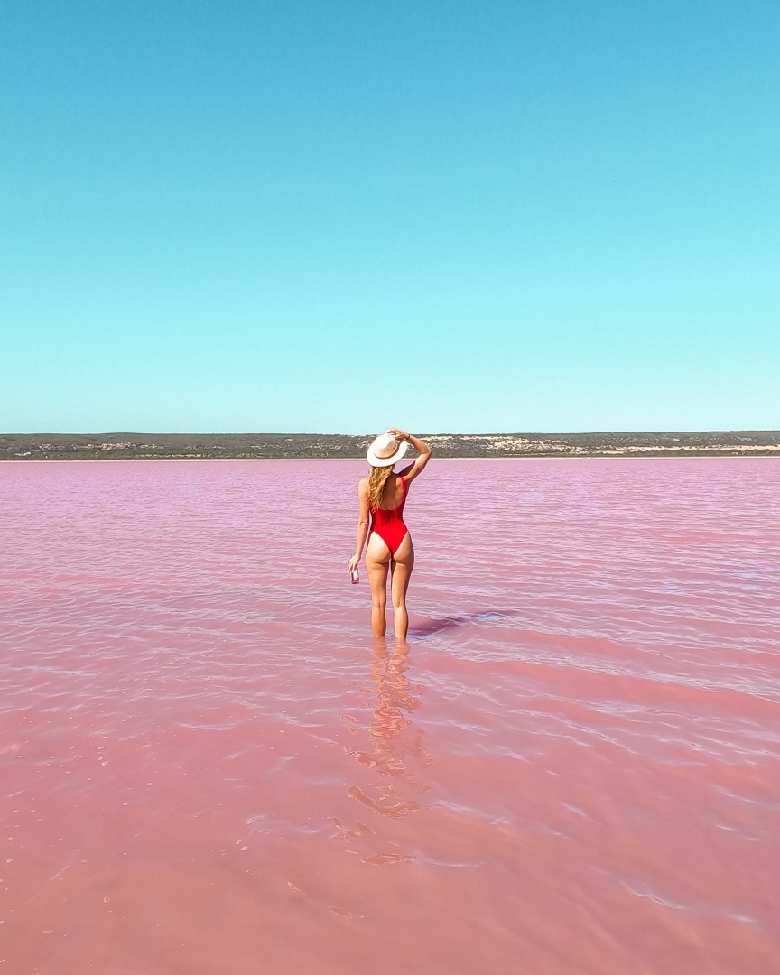 A woman in a red swimsuit and straw hat stands in the pink waters of Pink Lake, Hutt Lagoon in Kalbarri, Western Australia.