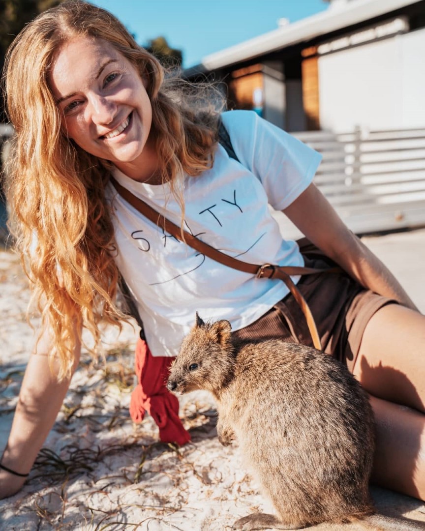 A woman with long blonde hair smiling at the camera while sitting next to a quokka on Rottnest Island Western Australia