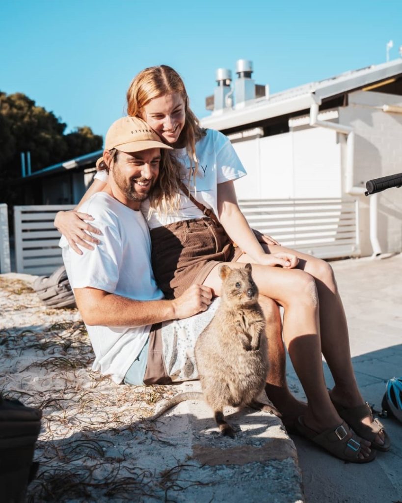 A couple sitting outdoors smiles as they both look at the quokka in front of them on Rottnest Island.