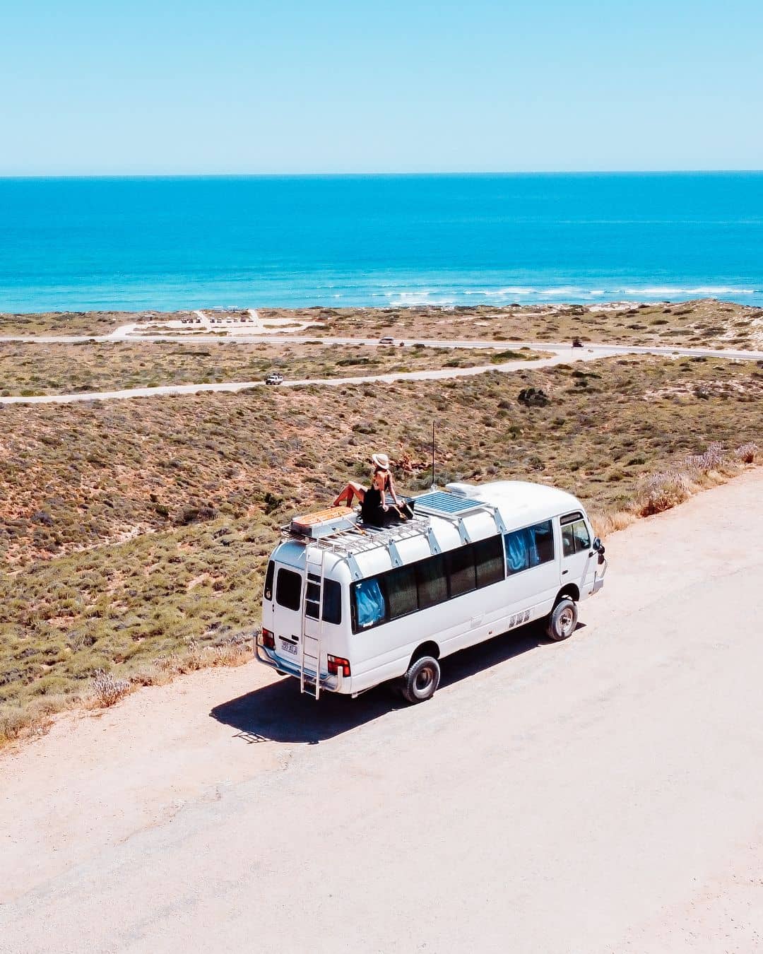 A person sits on the roof of a white camper van parked near the coastline in Cape Range National Park. The scene includes the blue ocean, sandy shoreline, and surrounding vegetation, creating a picturesque travel moment.