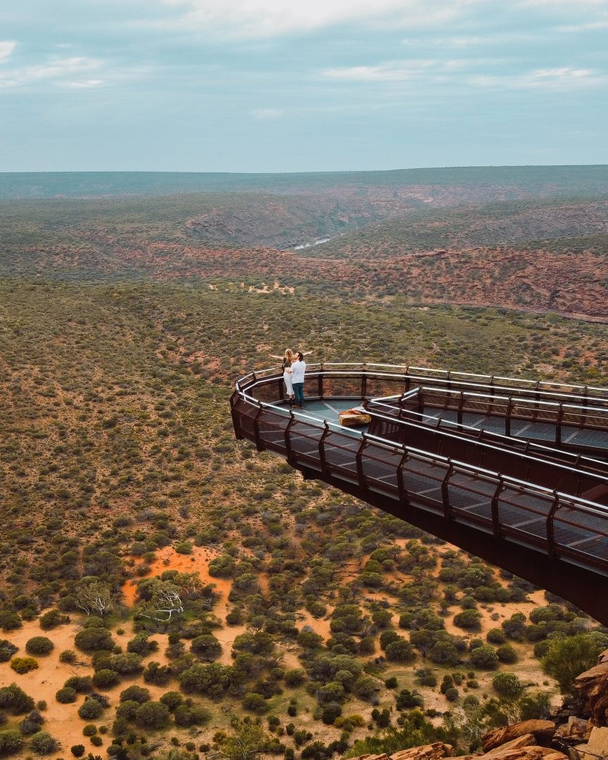 A couple stands on the Kalbarri Skywalk, a platform extending over the rugged landscape of Kalbarri National Park, Western Australia. The aerial view shows the vast, arid terrain with scattered greenery and winding gorges under a partly cloudy sky.