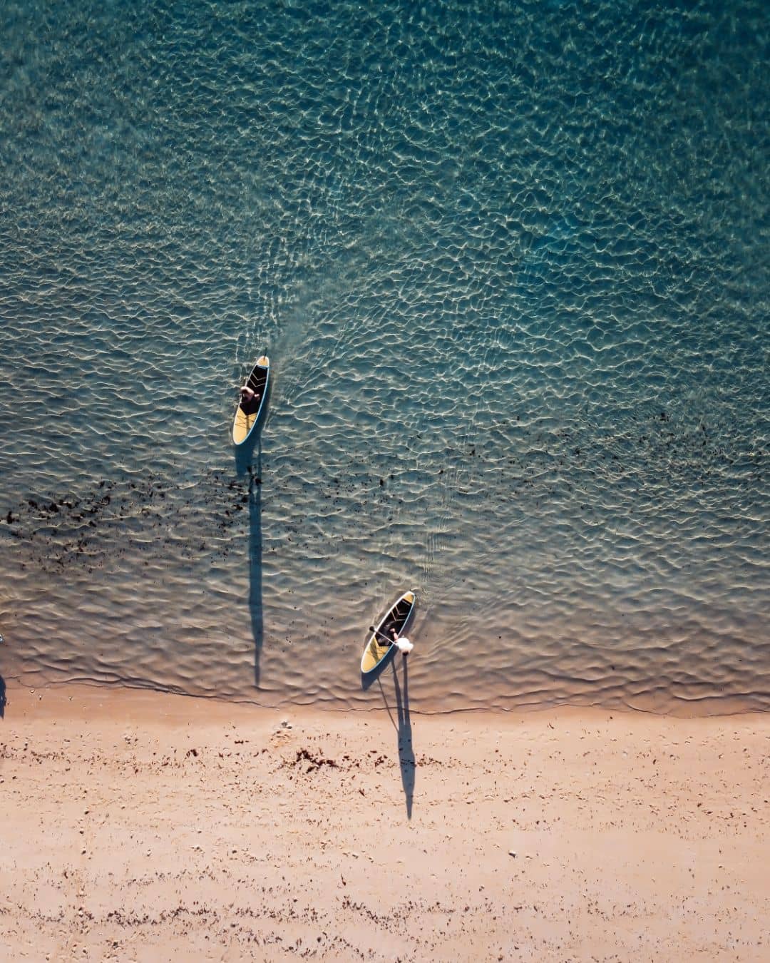 Aerial view of two kayaks pulled up on a sandy beach with clear, shallow water at South Lefroy near Coral Bay. The kayaks cast long shadows in the late afternoon sun.