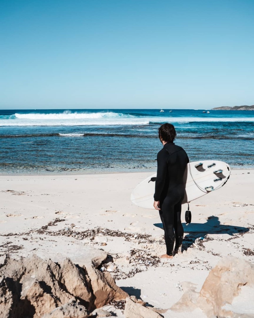 A surfer in a wetsuit holding a surfboard, standing on the sandy beach at Strickland Bay, Rottnest Island, looking out at the waves and the ocean.