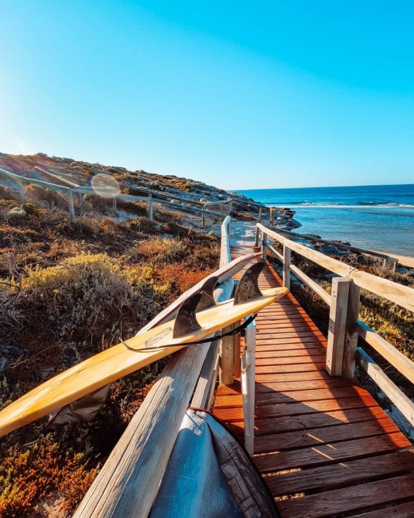 A surfboard resting on the railing of a wooden boardwalk leading down to the beach at Strickland Bay, Rottnest Island. The boardwalk is surrounded by coastal vegetation with the ocean visible in the background.