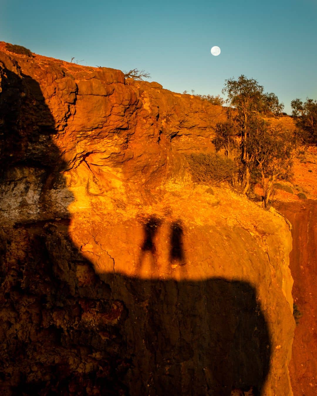 Two shadows are cast on a rocky cliff at sunset in Charles Knife Canyon, Cape Range National Park. The golden light illuminates the rugged landscape, with a full moon visible in the clear sky above.

