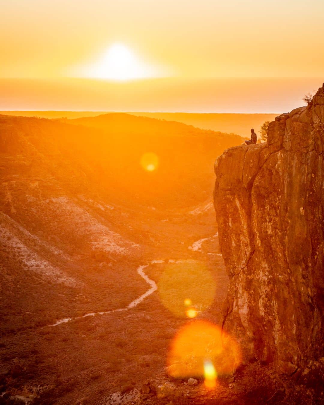 A person sits on a cliff at Charles Knife Canyon, Cape Range National Park, during sunset, with rugged terrain and winding paths below illuminated by golden hues.