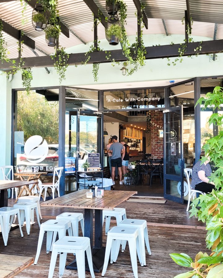 Entrance of "The Good Egg," a charming cafe located in Busselton, Western Australia. The exterior is decorated with lush hanging green plants, complementing the earthy wooden furniture. Inside, patrons can be seen ordering at the counter.