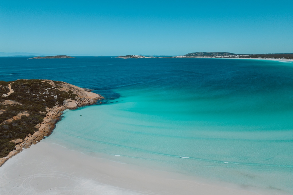 A scenic view of Wharton Beach in Esperance, a town near Bremer Bay in Western Australia. The shoreline is covered in soft white sand, curving around turquoise shallows that blend seamlessly into the deeper blue of the ocean. The rocky headlands and distant islands enhance the natural beauty.