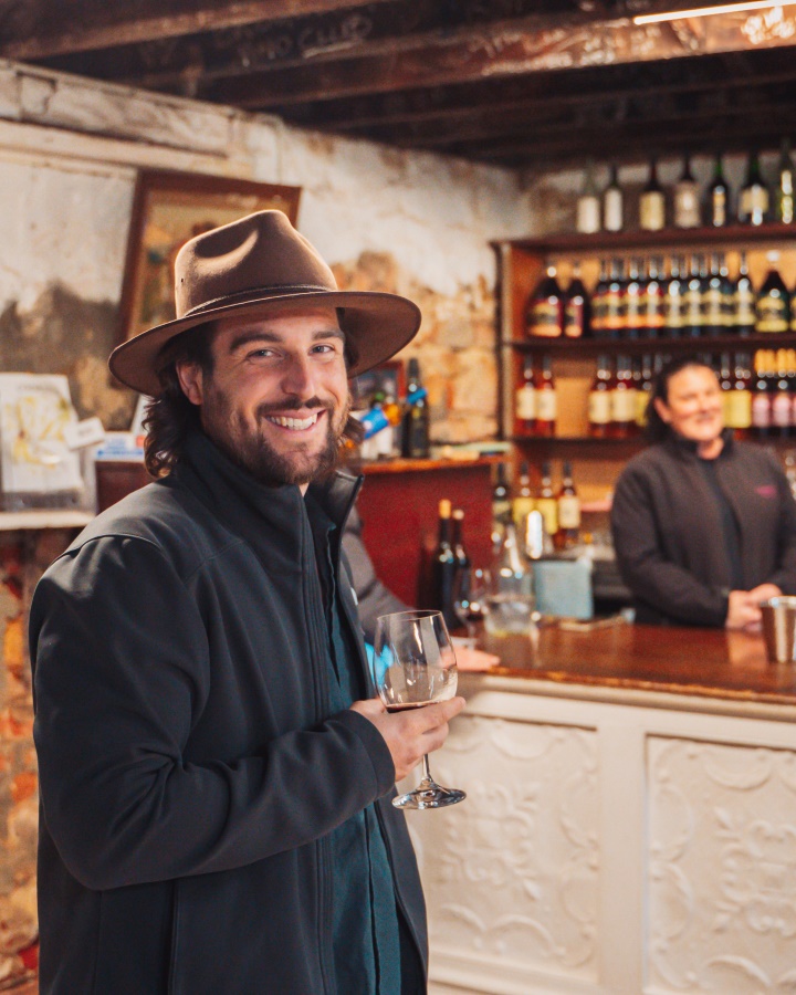 A man in a hat and jacket smiles while holding a glass of wine in a cozy, rustic wine cellar. It shows the charm of wine tasting in a traditional setting in Perth Western Australia