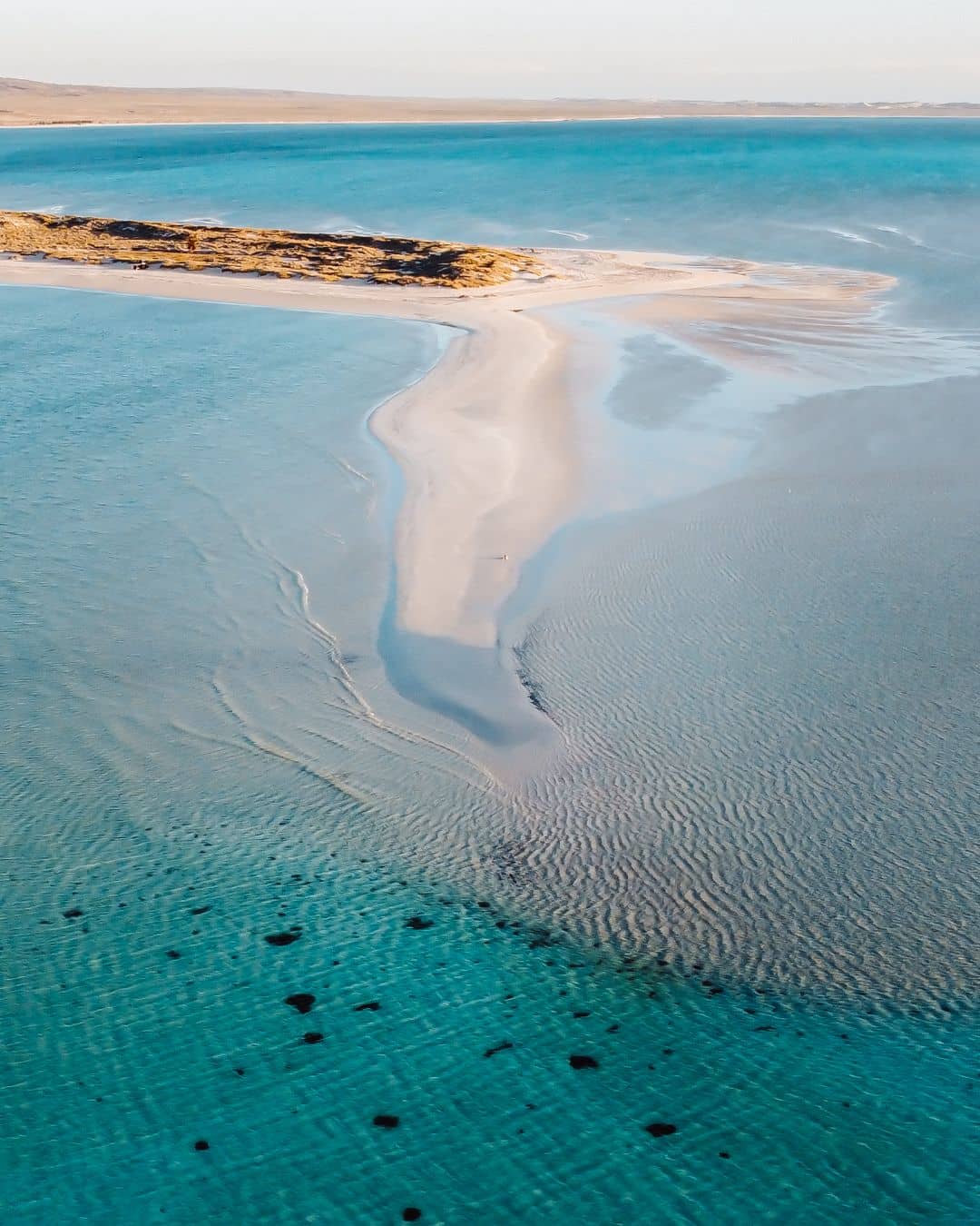 An aerial view of a sandbar extending into the turquoise waters near Coral Bay. The clear, shallow water reveals the sandy bottom, creating a beautiful gradient of blue hues. The sandbar leads to a small, rocky outcrop.