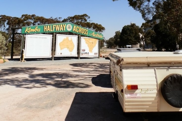 Sign of "Kimba, Halfway Across Australia" from a different angle, featuring a parked camper trailer in the foreground. The sign remains prominent in the background with its informational panels about Kimba and its halfway location across Australia.