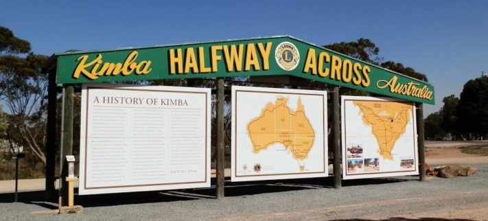 A large green and yellow sign in Kimba, South Australia, that reads "Kimba, Halfway Across Australia." Below it, there are three informational boards. The leftmost one provides a historical overview of Kimba, while the middle and right boards display maps illustrating its geographic location.