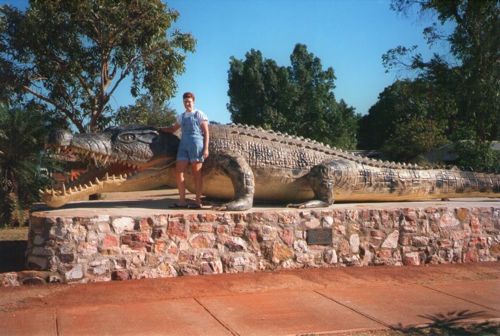 Woman in overalls standing in front of the biggest life-size crocodile replica in the world. It's a sunny day with clear skies and lush vegetation.