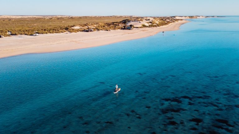 An aerial view of a tranquil beach scene in Exmouth, showcasing a wide expanse of clear blue waters bordered by a sandy beach. On the left, a few vehicles are parked on the dunes, while in the water, one person is standing on a paddleboard, alone in the sea. Perth to Exmouth tours