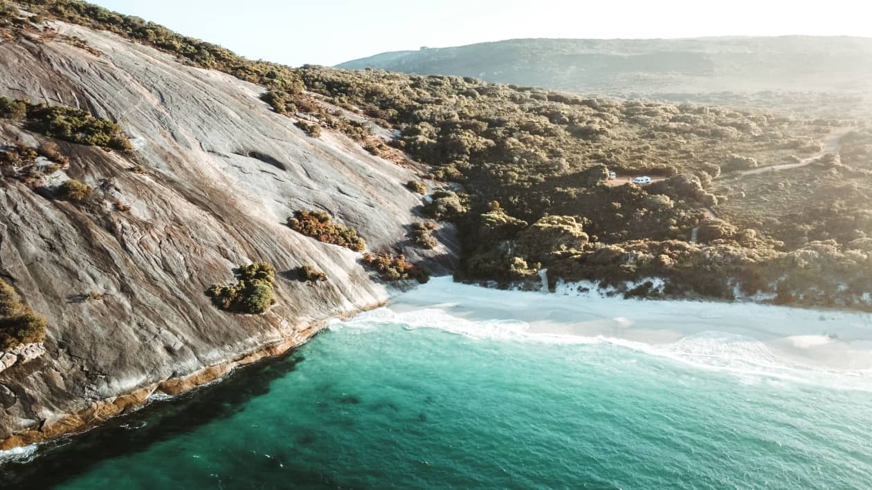 Aerial view of a secluded beach with turquoise waters, backed by a rocky slope and dense greenery near Augusta, Western Australia.