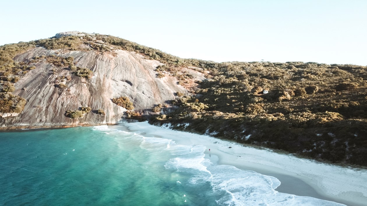A picturesque coastal scene featuring a rocky hill covered in vegetation beside a pristine sandy beach and turquoise waters in Augusta, Western Australia.