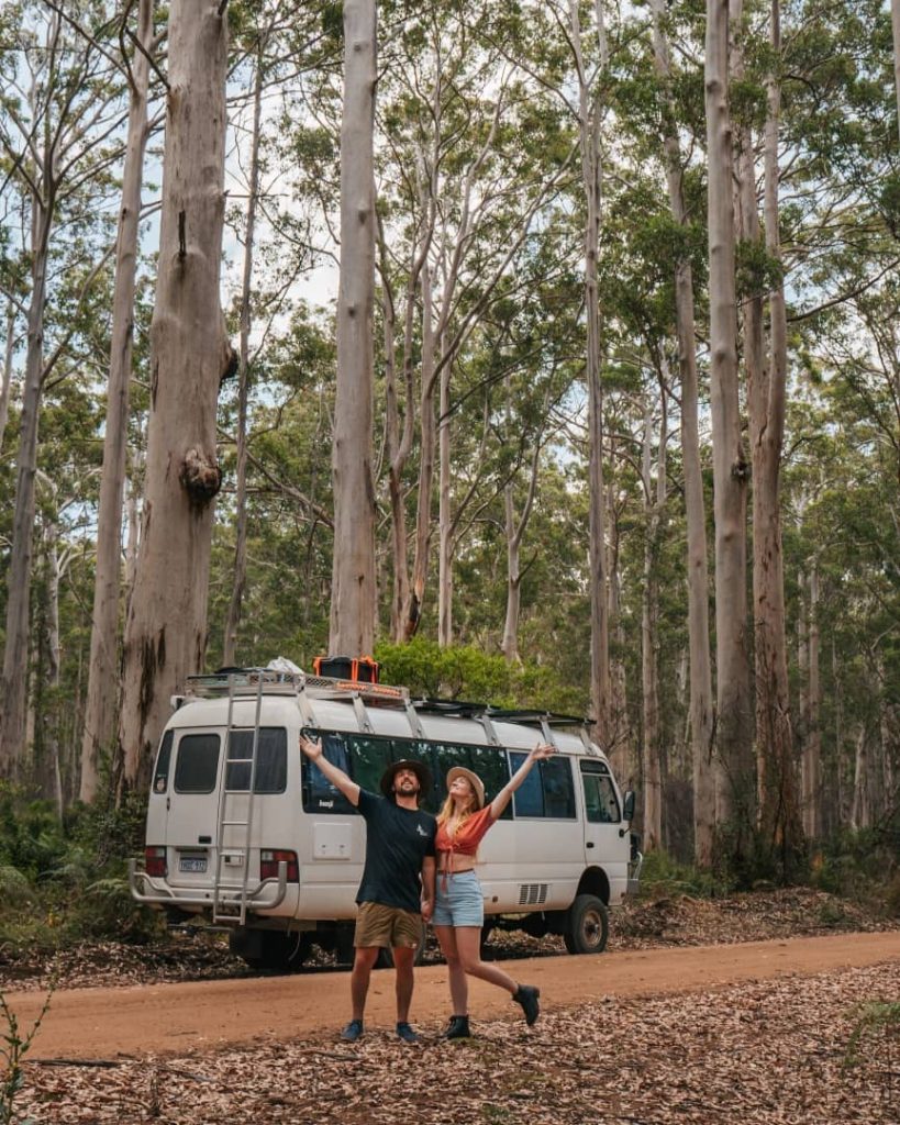 A couple stands joyfully in front of their camper van on a dirt road surrounded by tall trees in Boranup Forest, Margaret River, Western Australia.