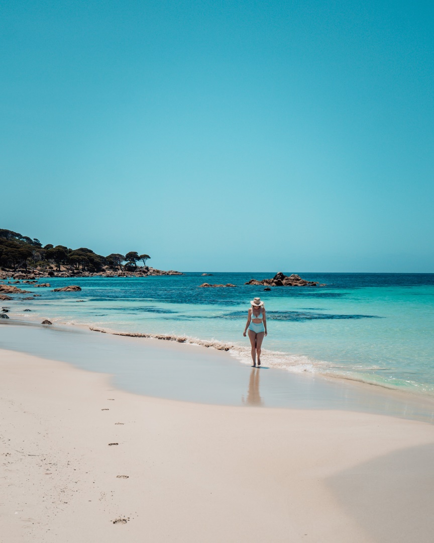 A woman walks along the pristine sandy shore of Bunker Bay Beach in Western Australia, with clear blue waters and rocky outcrops in the background. It is one of the best beaches in Margaret river region