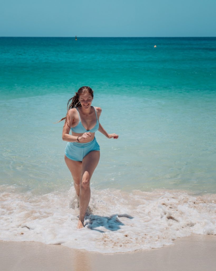 A woman in a blue bikini runs out of the turquoise ocean water onto the sandy beach of Bunker Bay, Western Australia
