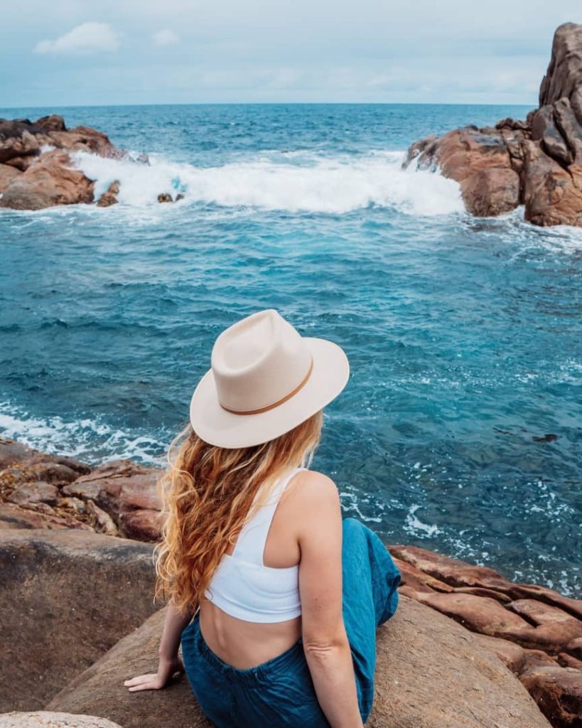 A woman in a hat sits on a rock, gazing at the waves crashing against rocky formations at Canal Rocks in Margaret River, Western Australia.