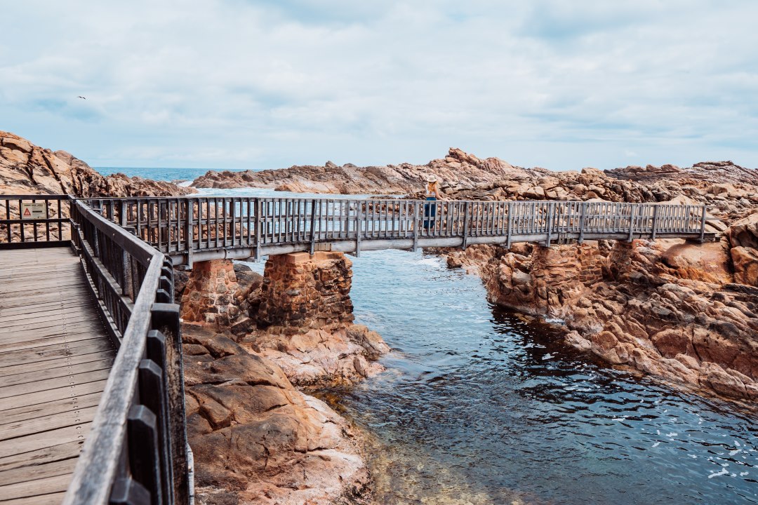 A wooden bridge stretches over a rocky coastline and clear waters at Canal Rocks in Margaret River, with a person standing on the bridge enjoying the scenic view. This image shows things to do in Pemberton in nearby places.