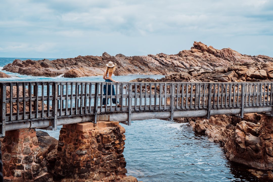 A woman walks across a wooden bridge over the water at Canal Rocks in Margaret River, Western Australia, with rocky formations in the background. The image shows things to do in Dunsborough