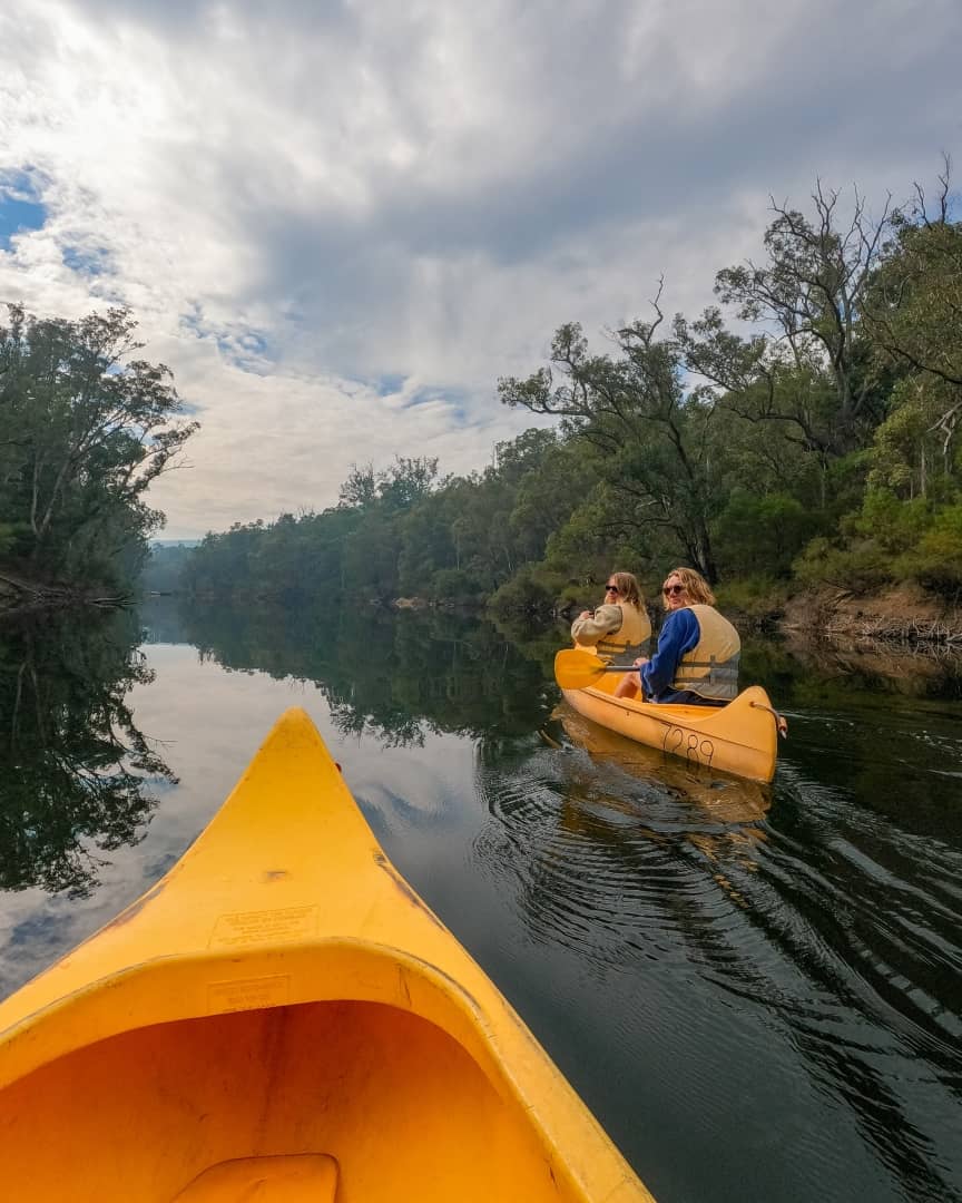 Two people paddle yellow canoes on a calm river surrounded by greenery and a partly cloudy sky showing things to do in Pemberton WA