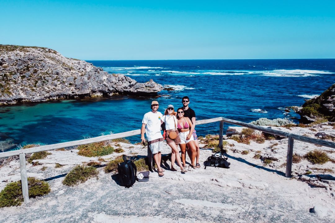 A group of four friends pose for a photo at Cape Vlamingh, Rottnest Island, with a stunning view of the rocky coastline and deep blue ocean behind them.