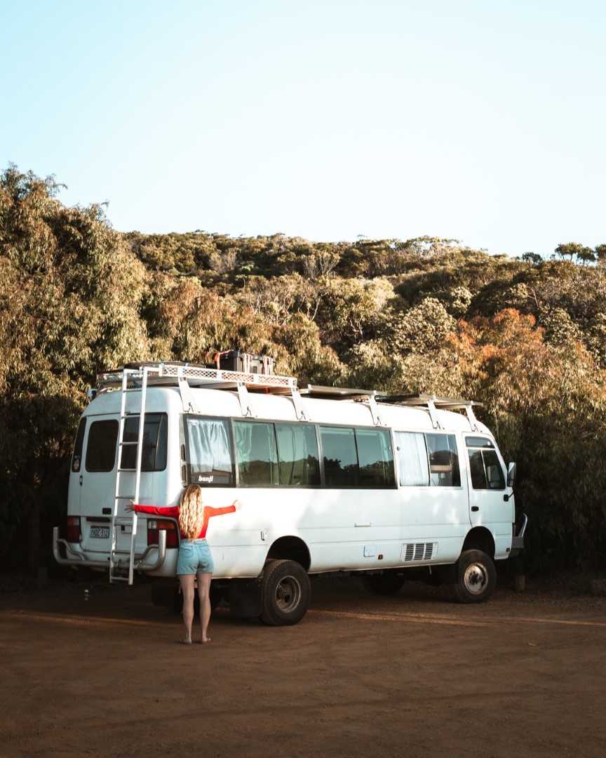 A woman hugs a white camper van parked on a dirt road, surrounded by lush vegetation in Augusta, Western Australia.