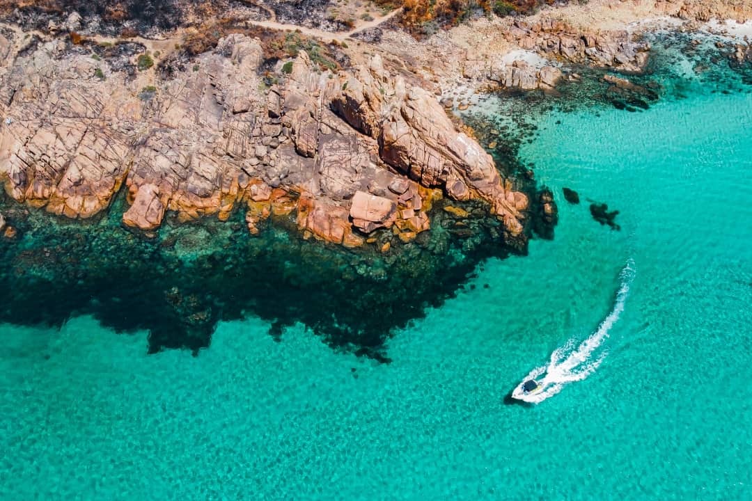 An aerial view of Castle Rock Bay in Western Australia shows turquoise waters, rugged orange and brown rock formations, and a boat speeding through the clear blue sea.