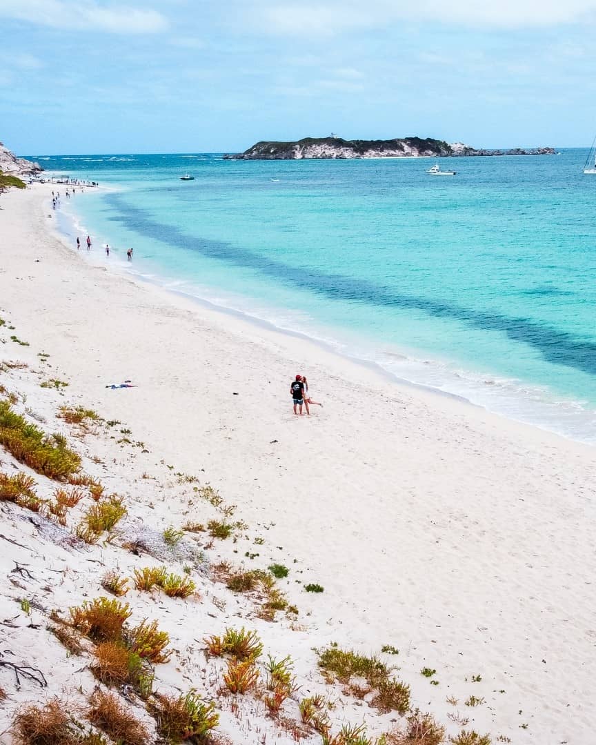A couple stands on the sandy beach of Hamelin Bay, Western Australia, with turquoise waters and a distant island in the background. It is one of the best beaches in Margaret River region