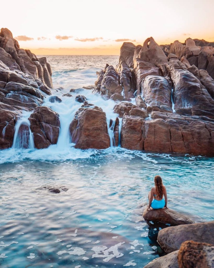 A woman in a blue swimsuit sits on a rock in the water at Injidup Natural Spa, Western Australia, with waves crashing through rocky formations at sunset.