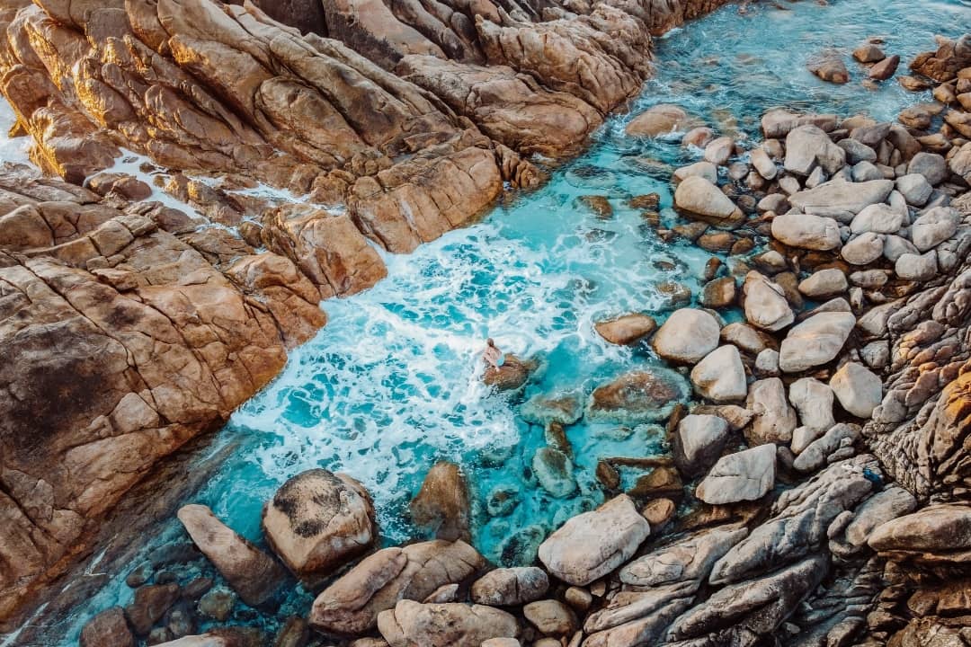 A person is sitting in the turquoise waters of the Injidup Natural Spa, Western Australia. The background shows rugged rocks and boulders.