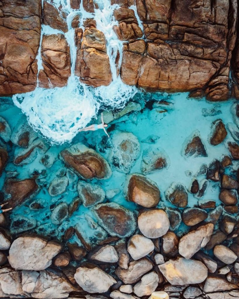 Aerial view of a person floating in the clear blue waters surrounded by rocks with waves cascading over the rocky formations in Injidup Natural Spa in Yallingup, Western Australia,