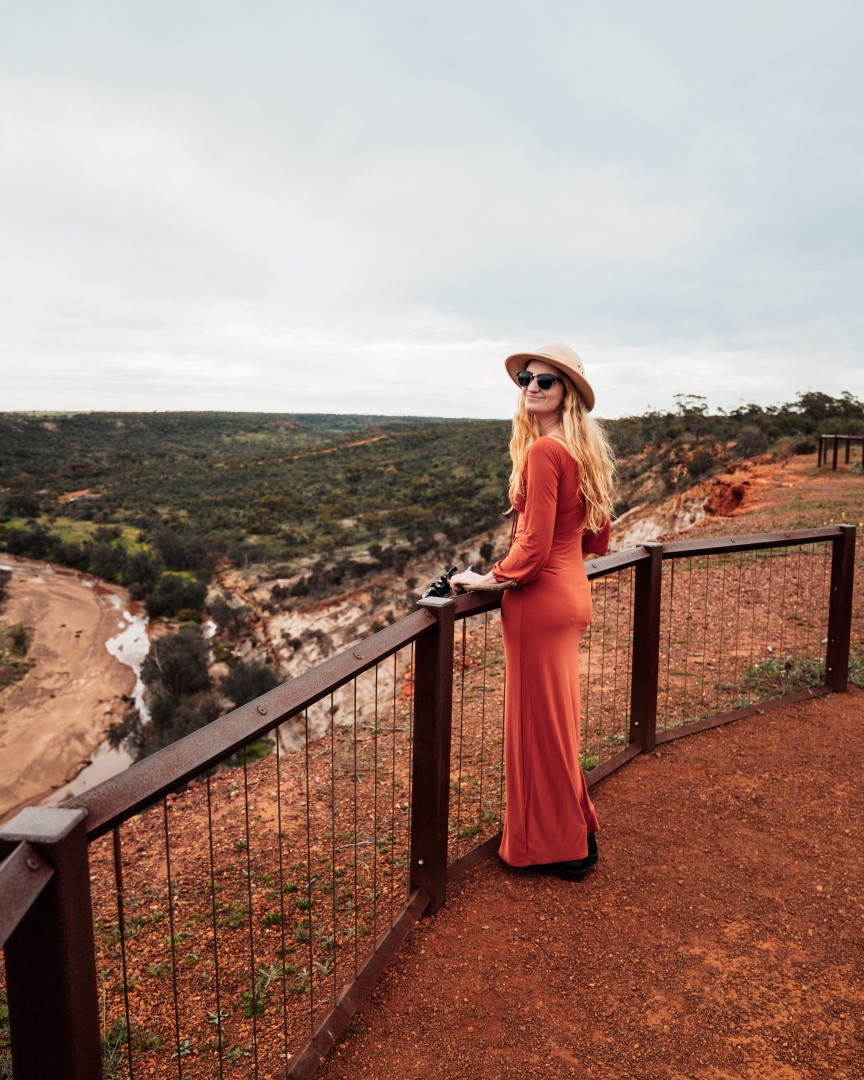 A woman in a long orange dress and hat stands at a lookout, gazing over the scenic landscape of Kalbarri, Western Australia.